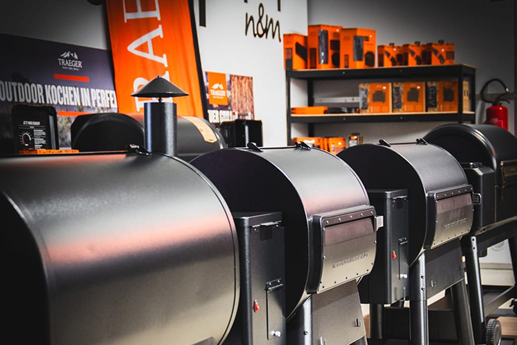 Eine Reihe schwarzer Traeger-Pelletgrills in einem Ausstellungsraum, im Hintergrund orangefarbene Banner und Regale mit Grillzubehör.