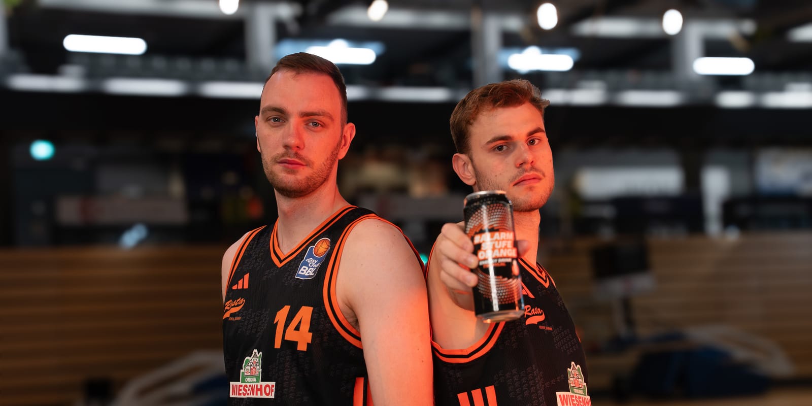 Zwei Basketballspieler in schwarzen und orangefarbenen Trikots posieren auf dem Spielfeld; einer hält eine Dose in Richtung Kamera.