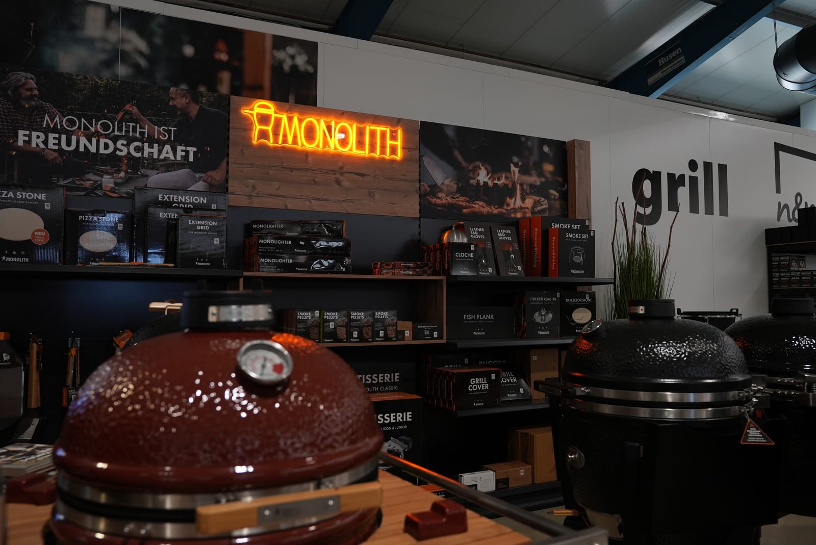 Ein Stand, an dem Monolith-Kamado-Grills mit diversem Zubehör und einer Leuchtreklame ausgestellt sind, mit Regalen voller Grillprodukte und heller Beleuchtung.