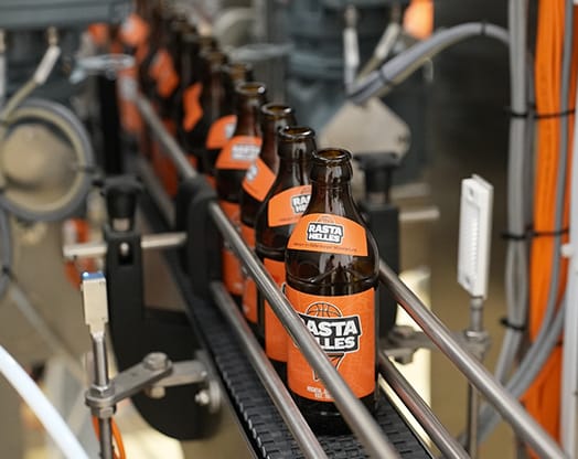 Förderband mit braunen Bierflaschen mit „Rasta Helles”-Etiketten in einer Brauerei mit Maschinen im Hintergrund.
