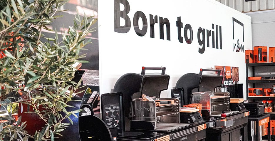 Ausstellung von Grillgeräten mit dem Schild „Born to grill“ darüber, umgeben von Grillzubehör und einer Topfpflanze.