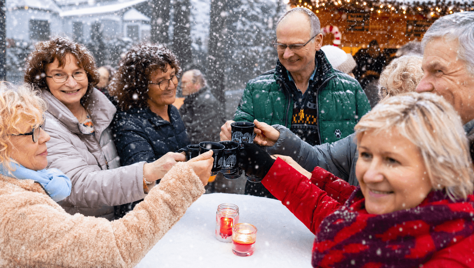 Eine Gruppe Menschen steht um einen Tisch und stoßen mit Tassen an. Die Atmosphäre ist weihnachtlich und es fällt Schnee.