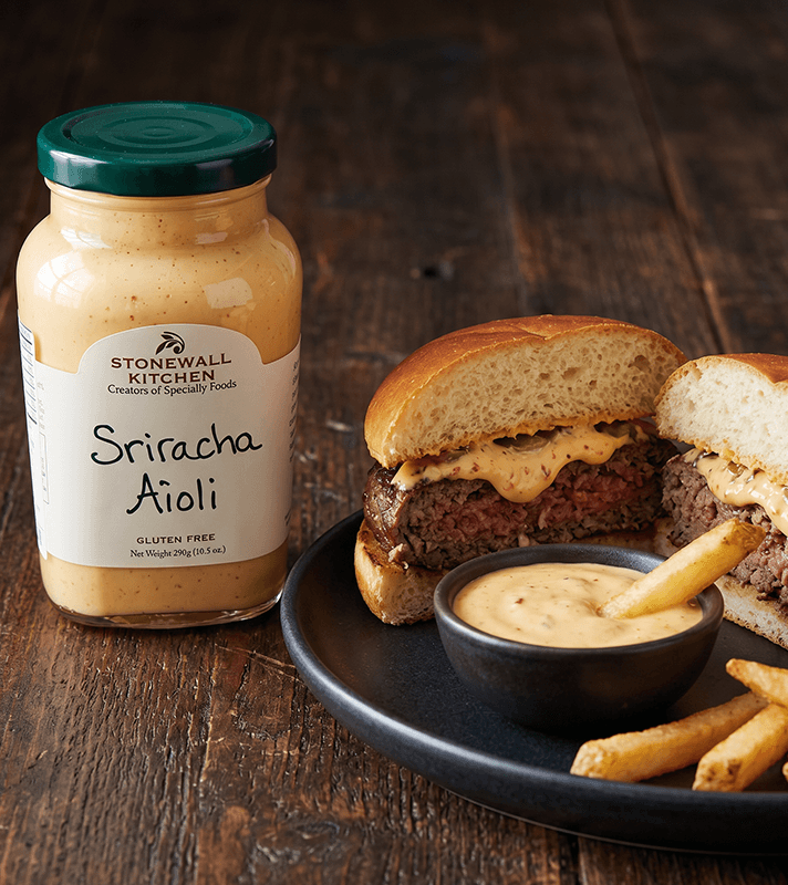 Glas Sriracha Aioli neben einem aufgeschnittenen Burger mit Aioli, Pommes Frites und einer Schale Aioli auf dunklem Holztisch.