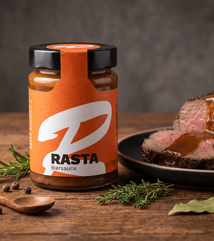 Glas mit RASTA-Biersauce (300 g) auf einem rustikalen Holztisch. Im Hintergrund liegt auf einem schwarzen Teller rosa gebratenes Rindfleisch mit brauner RASTA Biersauce. Frische Kräuter, Pfefferkörner und ein Holzlöffel ergänzen die Szene und unterstreichen den herzhaften Genusscharakter des Produkts.