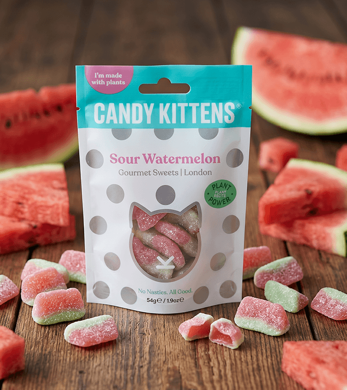 Tüte Candy Kittens "Sour Watermelon" mit Katzenfenster, umgeben von Wassermelonenstücken und Gummibärchen auf Holz.