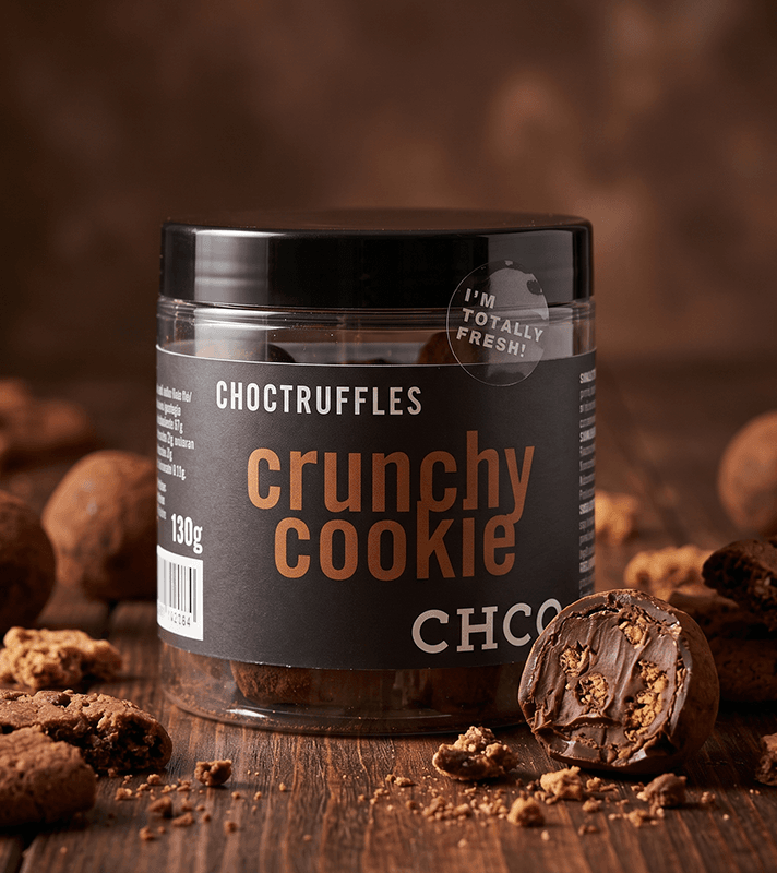 Transparenter Behälter mit "Chocotruffles Crunchy Cookie" Aufschrift, umgeben von Schokoladentrüffeln und Keksstücken auf Holz.