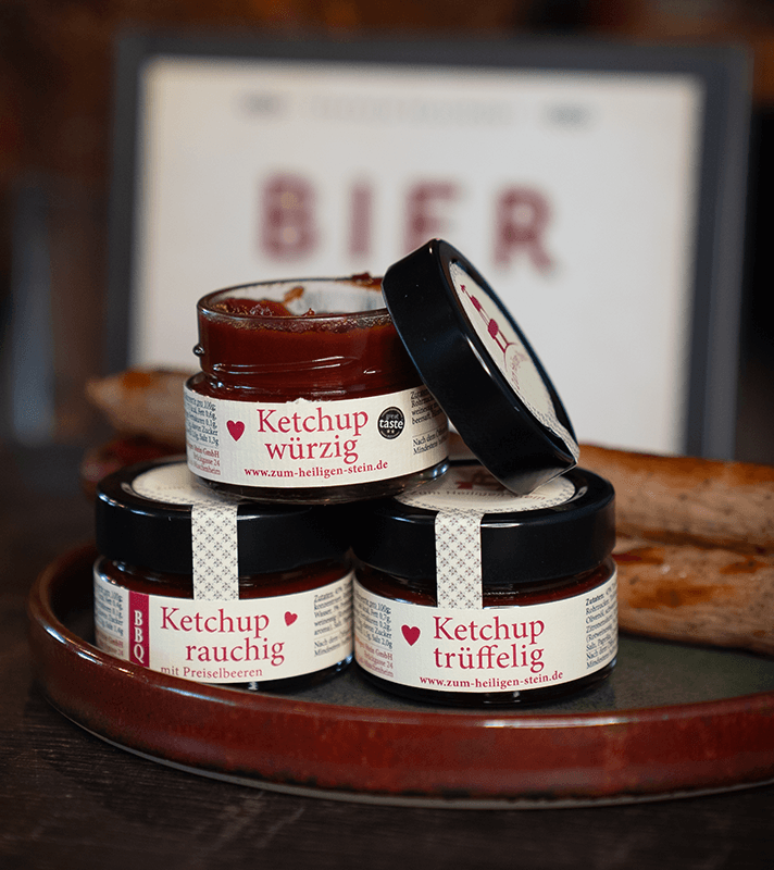 Drei Gläser mit würzigem, rauchigem und trüffeligem Ketchup auf einem Teller, mit schwarzem Deckel und weißen Etiketten, im Hintergrund ein Bildschirm mit "Bier".
