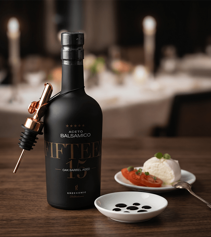 Schwarze Balsamico-Flasche mit Ausgießer, daneben eine Schale mit Balsamico-Tropfen, Mozzarella und Tomaten auf Holzoberfläche.