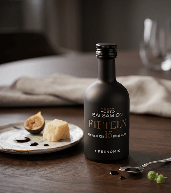 Schwarze Flasche Aceto Balsamico "Fifteen" mit goldenem Etikett, daneben Teller mit Feige, Käse und Balsamico-Tropfen, Löffel mit Basilikum.