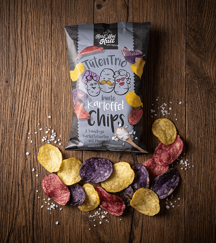 Bunte Kartoffelchips in lila, gelb und rot, umgeben von Salz, vor einer schwarzen Verpackung mit buntem Design und Cartoon-Illustrationen auf Holzuntergrund.
