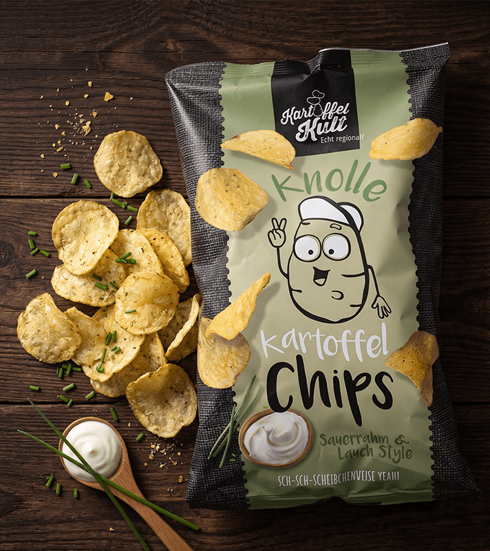 Bunte Kartoffelchips in einer grünen und schwarzen Verpackung mit Cartoon-Kartoffel, daneben auf Holzuntergrund, einige Chips und ein Löffel mit Sauerrahm.