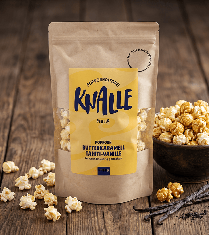 Braune Papiertüte mit gelbem Etikett "Knalle Popkorn Butterkaramell Tahiti-Vanille", daneben Schale mit Popcorn, Vanilleschoten.