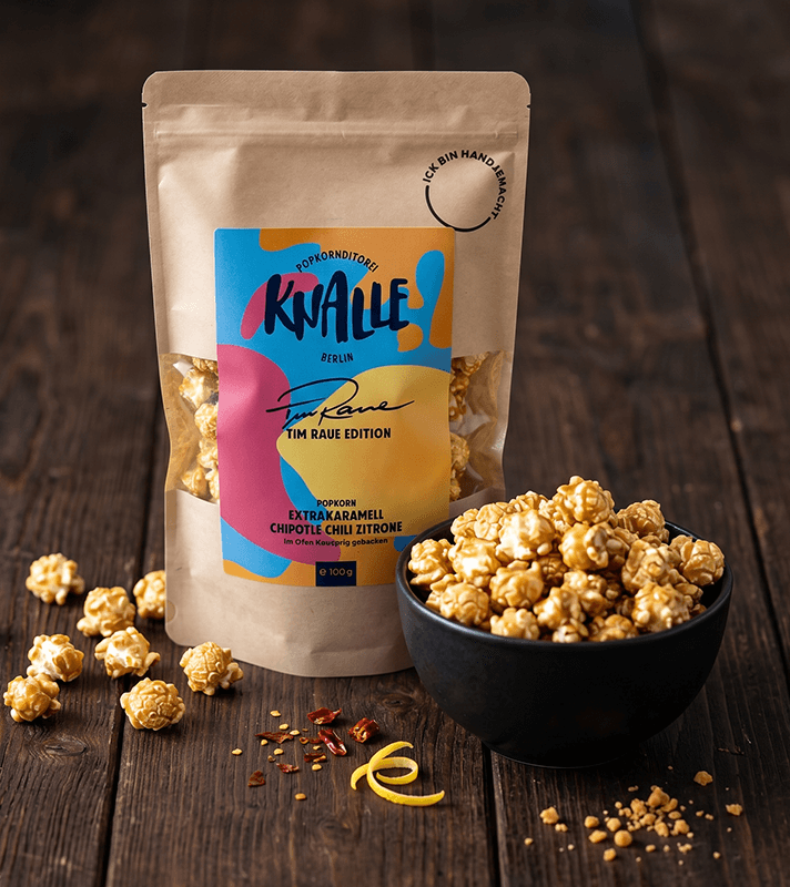 Popcorn-Tüte „Knalle“ mit Karamell-Chili-Zitronen-Popcorn in Schale und verstreut, dazu Chiliflocken und Zitronenschale auf Holz.