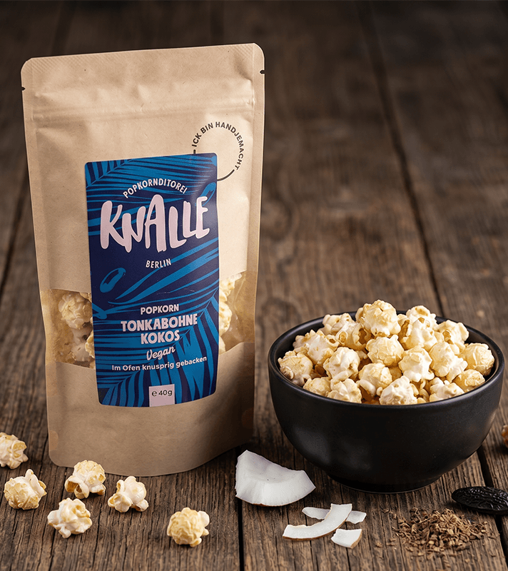 Popcorn-Beutel "Knalle Tonkabohne Kokos" neben schwarzer Schale mit Popcorn, Kokosstücken und Tonkabohnen auf Holz.