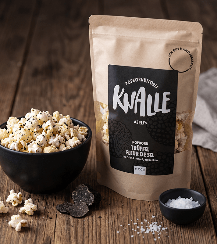 Popcorn Trüffel Fleur de Sel in einer schwarzen Schale und in einer braunen Papiertüte, daneben Trüffelstücke und Meersalz.