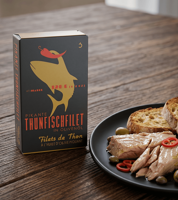 Schwarze Verpackung "Pikante Thunfischfilet in Olivenöl" mit goldenem Thunfisch-Logo und roter Chili, daneben Teller mit Thunfisch, Brot, Oliven.