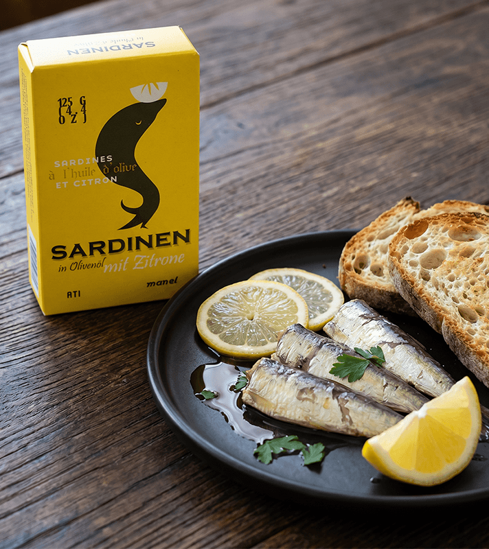 Gelbe Sardinendose mit schwarzem Sardinen-Logo, daneben schwarzer Teller mit 3 Sardinen, Zitronenscheiben, Petersilie und 2 Brotscheiben.