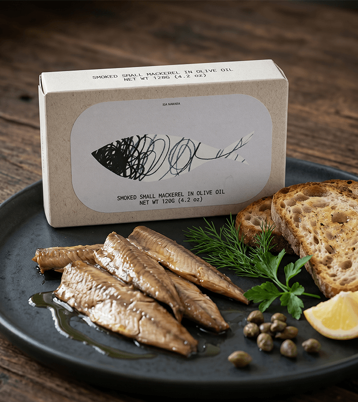 Geräucherte Makrelenfilets in Öl auf schwarzem Teller mit Kapern, Zitrone, Brot und Kräutern, dahinter eine Verpackung.