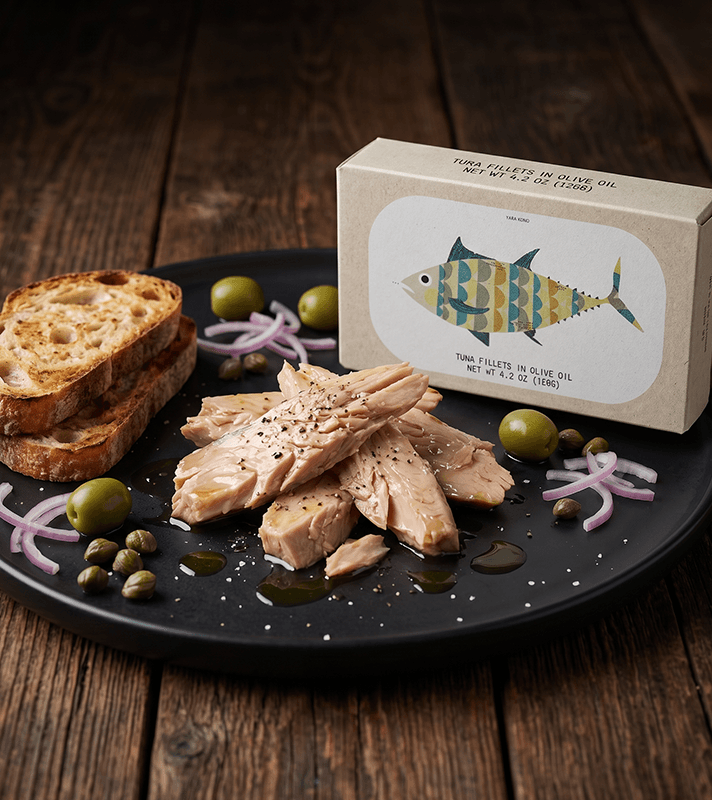 Thunfischfilets in Olivenöl auf schwarzem Teller mit geröstetem Brot, Oliven, Zwiebeln, Kapern und einer Verpackung mit Fischmotiv.