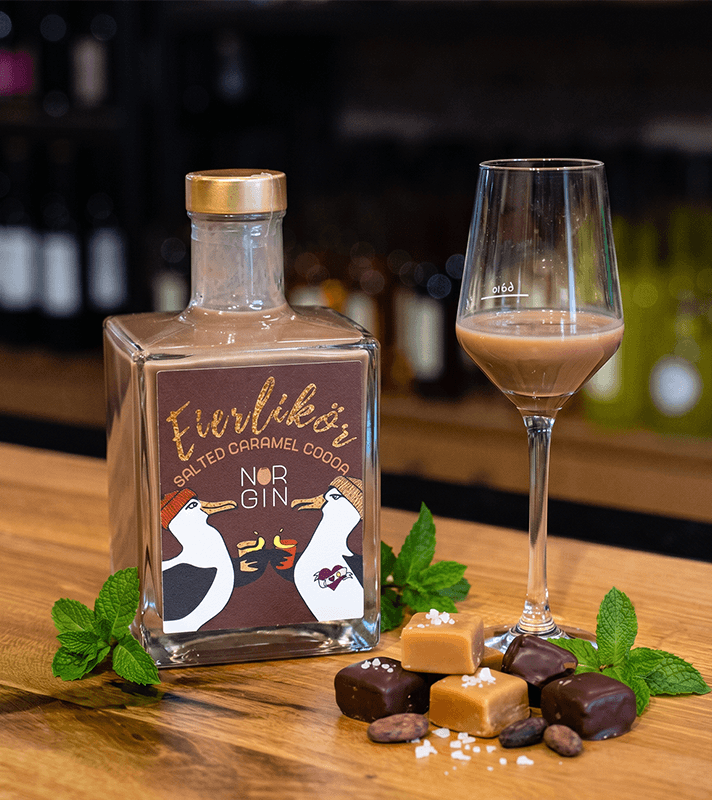 Flasche Everlikör Salted Caramel Cocoa Nor Gin mit Pinguin-Etikett, daneben ein Glas mit Likör, Minze, Karamell, Schokolade und Kakaobohnen.