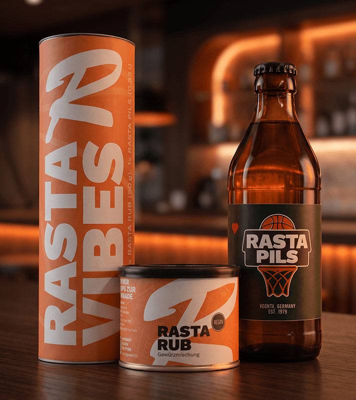 RASTA Vibes Geschenkset mit einer Flasche RASTA Pils (0,33 l), einer Dose RASTA Rub Gewürzmischung (90 g) und einer orangefarbenen Geschenkverpackung mit großem RASTA-Schriftzug. Das Set steht auf einer Holztheke vor einem stimmungsvoll beleuchteten Barhintergrund und eignet sich ideal als Geschenk für Grill- und Basketballfans.