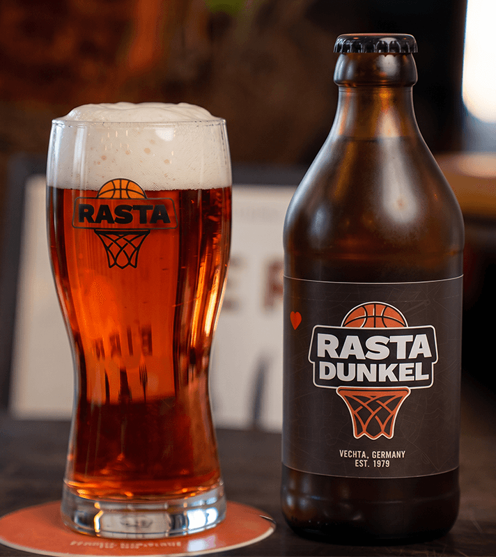Eine braune Bierflasche mit Rasta Dunkel-Logo, daneben ein gefülltes Bierglas mit Schaumkrone und Rasta Dunkel-Logo, auf einem Holztablett.