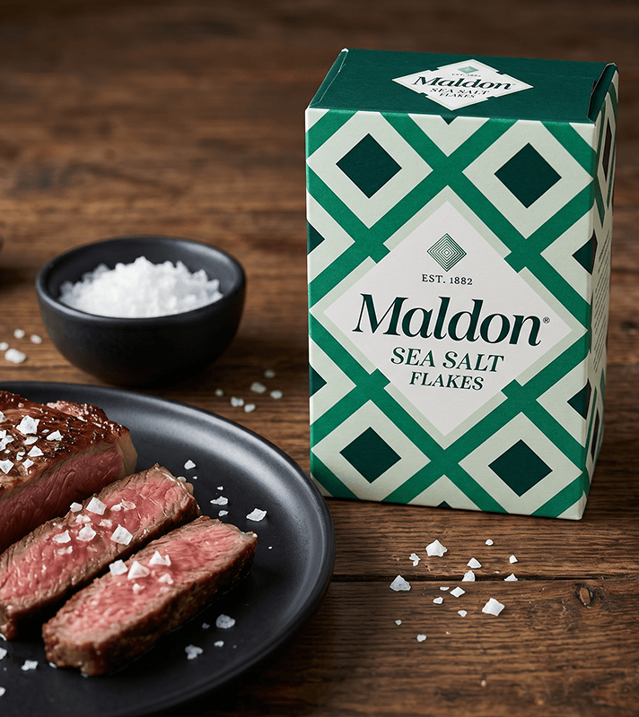 Maldon Meersalzflocken-Packung neben einem Teller mit geschnittenem Steak, bestreut mit Salzflocken, und einer kleinen Schale mit Salz.
