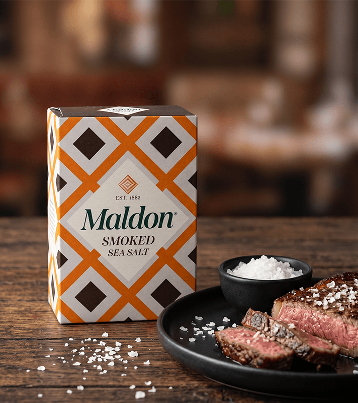 Maldon Smoked Sea Salt Packung neben Steak auf Teller mit Salzschale, Holzuntergrund.