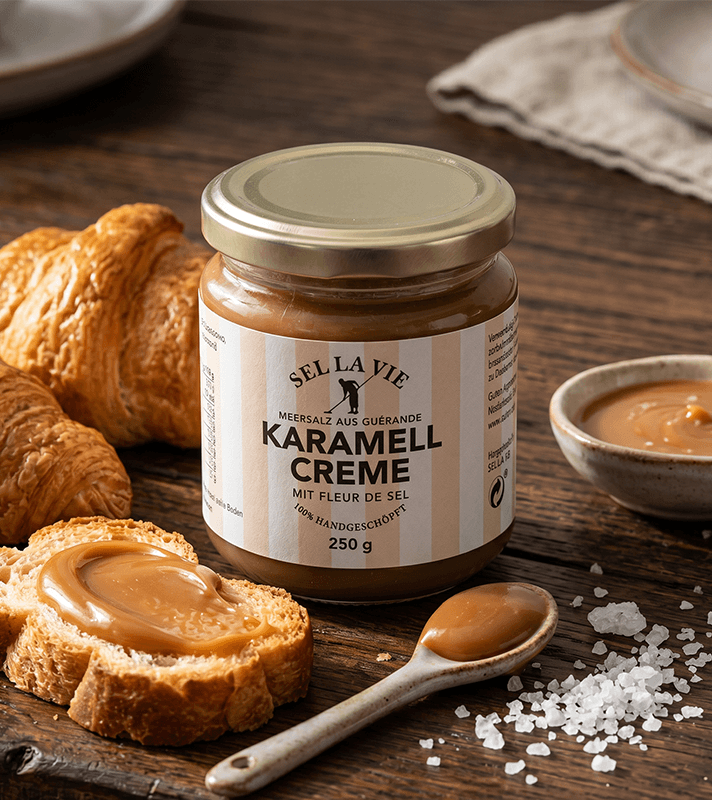 Ein Glas Karamellcreme mit Fleur de Sel, daneben Croissants, geröstetes Brot mit Creme, eine Schale und ein Löffel mit Creme, Meersalz.