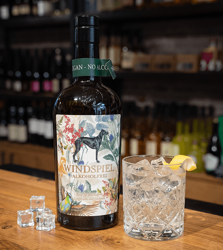Dunkle Flasche "WIND SPIEL ALKOHOLFREI" mit buntem Etikett (Hund, Vögel, Blumen), daneben Glas mit Eis, Zitrone und Eiswürfel.