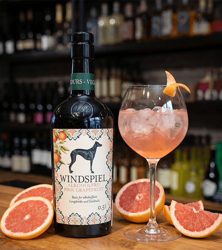Flasche Windspiel alkoholfrei Pink Grapefruit mit Etikett, daneben ein Glas mit rosa Getränk, Eiswürfeln und Grapefruit-Zeste, umgeben von Grapefruit-Scheiben.