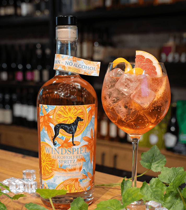 Flasche Windspiel alkoholfreier Spritz mit Hundemotiv, daneben ein Glas mit Eis, Grapefruit und Orangenschale, im Hintergrund Bar.