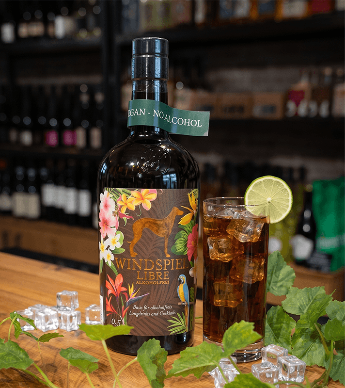 Braune Flasche "WIND SPIEL LIBRE ALKOHOLFREI" mit tropischem Etikett, daneben ein Glas mit braunem Getränk, Eis und Limettenscheibe.